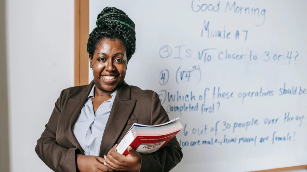 professora de pele negra segurando livro em frente a quadro branco com anotações